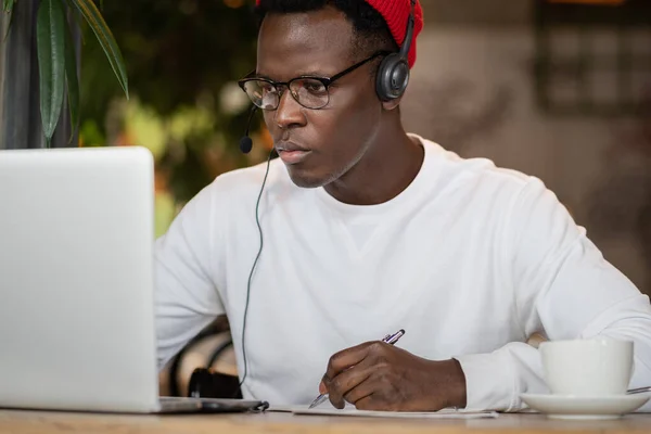 Afro man wear wear headphones, remotely online work in cafe, makes notes, looking webinar.