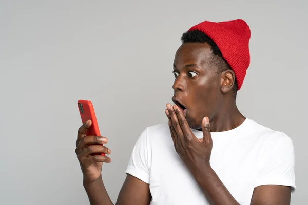 Überraschter Afro-Mann macht große Augen auf, bedeckt ihren Mund schockiert von Nachricht, die auf Handy schaut — Stockfoto