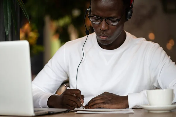 Afro homme porter des écouteurs, travail à distance en ligne dans le café, prend des notes, écouter des cours en ligne. — Photo