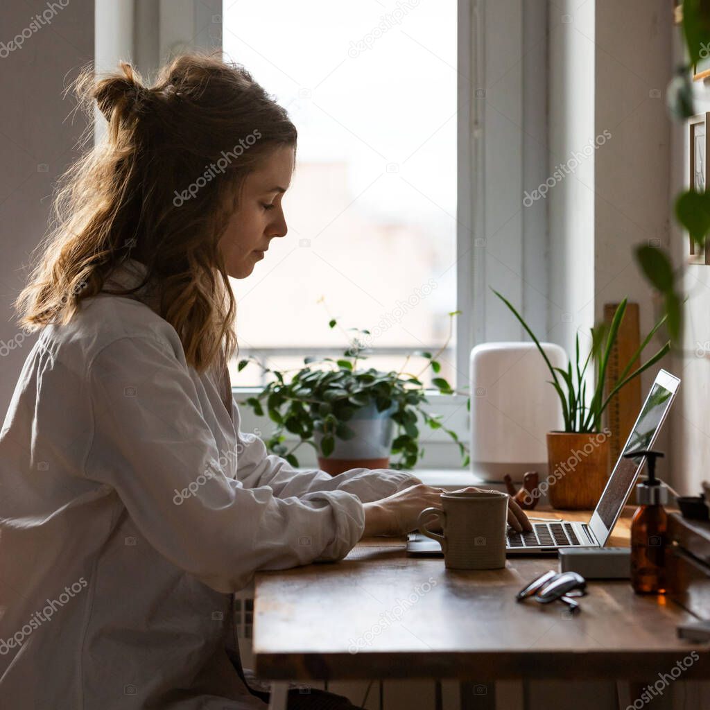 Young woman freelancer/designer working on computer from home office during self-isolation due to coronavirus. Cozy workplace surrounded by plants. Remote work, Telecommuting, Distance job.