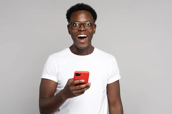 Glücklicher afrikanischer Millennial-Mann mit Brille trägt weißes T-Shirt, Handy in der Hand, isoliert im Studio — Stockfoto