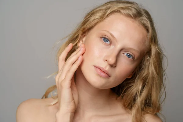 Ung kvinna ansikte med blå ögon, lockigt naturligt blont hår, har ingen makeup, vidrör hennes mjuka hud — Stockfoto