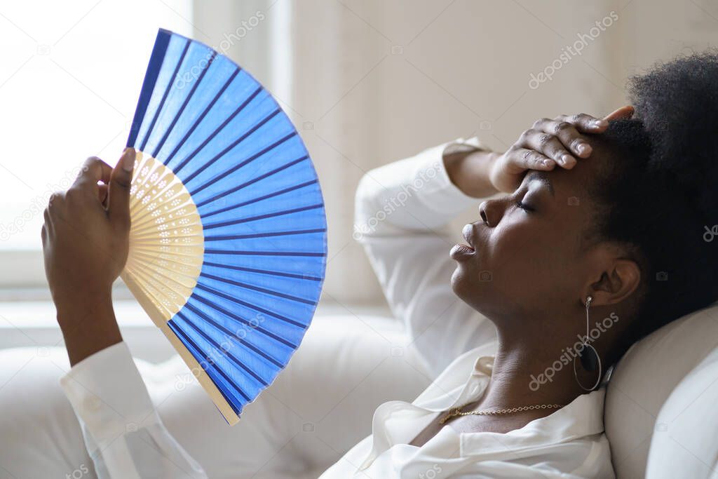 Afro business woman suffering from heat stroke sitting in living room at home using waving fan
