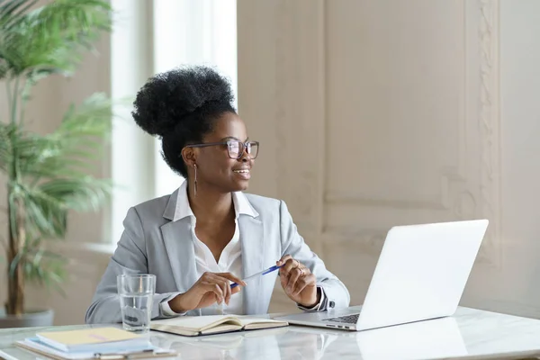 Sorrindo afro mulher em blazer usar óculos trabalhando no computador portátil em casa escritório, olhando para longe. — Fotografia de Stock