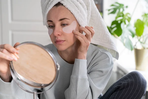 Mulher com toalha na cabeça aplicar hidrogel sob os olhos remendos de recuperação em casa, olhando no espelho. — Fotografia de Stock