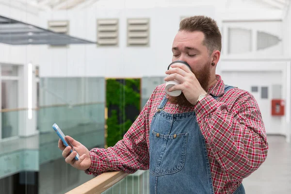 ブルートベアードの自営業担当者は ブルーオーバーオール チェックシャツ 紙コップからコーヒーを飲む スマートフォンを使用しています 休憩する時間だ ヒップスター床屋の男が休んでる 仕事中の休憩 — ストック写真