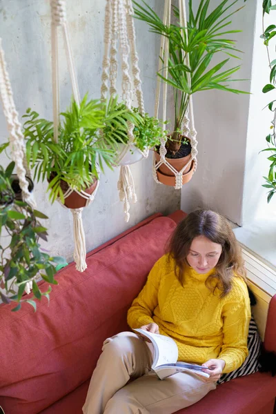 Mujer descansando, sentada en el sofá debajo de la percha de macramé de algodón con plantas de interior, revista de lectura —  Fotos de Stock