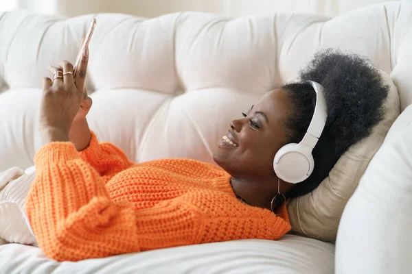 Femme africaine en pull porter des écouteurs, écouter de la musique, en utilisant un téléphone intelligent, couché sur le canapé à la maison — Photo