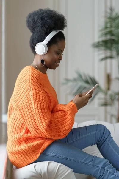 Femme millénaire noire en pull orange porter écouteurs sans fil écouter de la musique, en utilisant un téléphone intelligent — Photo