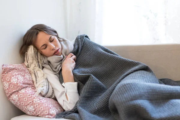 病気の若い女性がソファに横たわって震え カバーレットやスカーフに包まれ インフルエンザの症状を持って 自分自身を暖めようとします — ストック写真