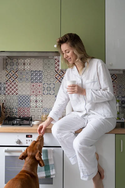 Kvinna i pyjamas håller vit mugg, dricka te på vintern morgon hemma behandlar hund med godsaker — Stockfoto