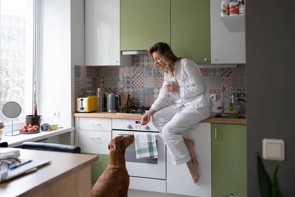 Woman in pajamas sitting in the kitchen, holding white mug, treats her beloved dog with goodies — Stock Photo, Image