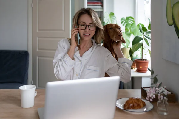 Woman in sleepwear sitting in living room, hugging her dog, taking on mobile phone working at laptop — Stock Photo, Image