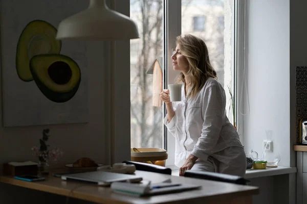 Pensive kvinna sitter på fönsterbräda, tittar genom fönstret dricka te på morgonen hemma. — Stockfoto