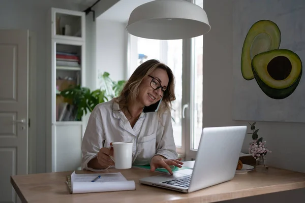 Woman sitting at home in the morning, drinking tea, taking on mobile phone, remote working at laptop — Stock Photo, Image