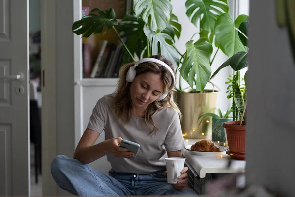 Kvinna lyssnar på musik bära hörlurar med hjälp av smarttelefon chatta i sociala nätverk, dricka te — Stockfoto