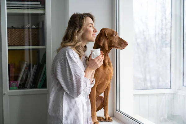 Propriétaire heureuse femme et chien assis sur le rebord de la fenêtre, tenant une tasse de café, regardant par la fenêtre — Photo