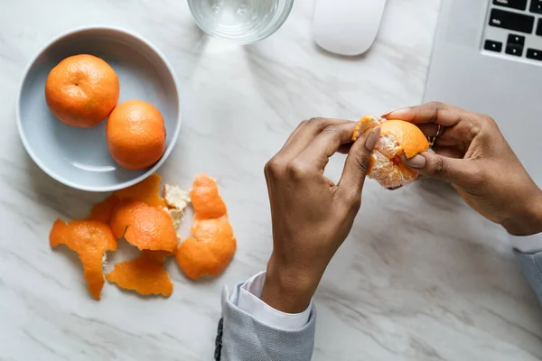 Afro alkalmazott nő kéz peeling érett édes mandarin, visel blézer, ül az asztalnál a munkahelyen — Stock Fotó