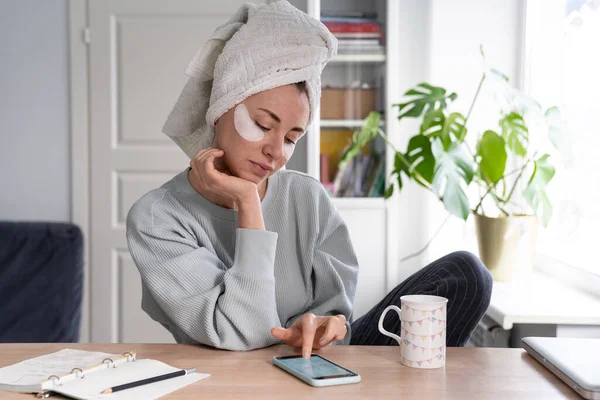 Mulher com toalha na cabeça e manchas sob os olhos usando telefone inteligente, lendo redes sociais alimentar — Fotografia de Stock