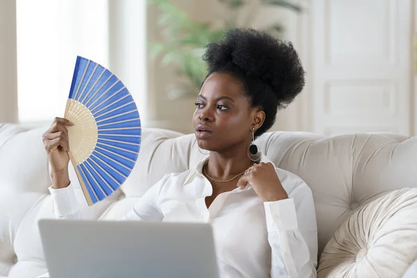 Afro mulher de negócios que sofre de insolação sentado na sala de estar em casa usando ventilador de acenar — Fotografia de Stock