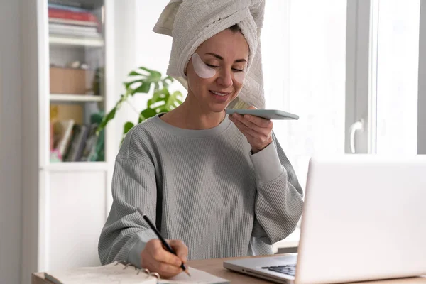 Mulher com toalha na cabeça remendos sob os olhos trabalho de casa no laptop registros mensagem de voz no celular — Fotografia de Stock