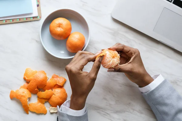 Afro alkalmazott nő kéz peeling érett édes mandarin, visel blézer, ül az asztalnál a munkahelyen — Stock Fotó