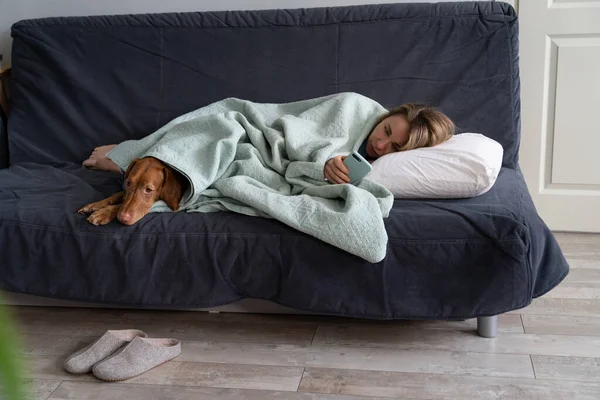 Smutná žena ležící na pohovce se psem, pomocí chytrého telefonu čekat na přítele volání nebo SMS, cítí depresi — Stock fotografie