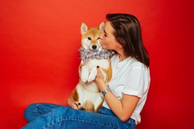 Beyaz tişörtlü, kot pantolonlu, gümüş madalyalı, kırmızı arka planda Shiba Inu köpeğini öpen tatlı esmer kadın. Hayvanlara sevgi, evcil hayvan konsepti