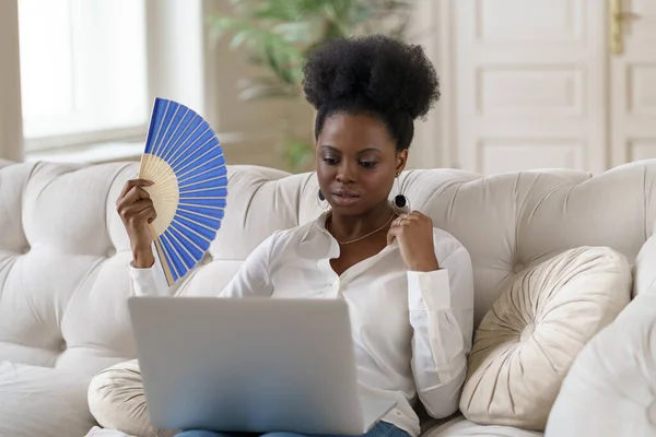 Afro mulher de negócios que sofre de insolação sentado na sala de estar em casa usando ventilador de acenar — Fotografia de Stock