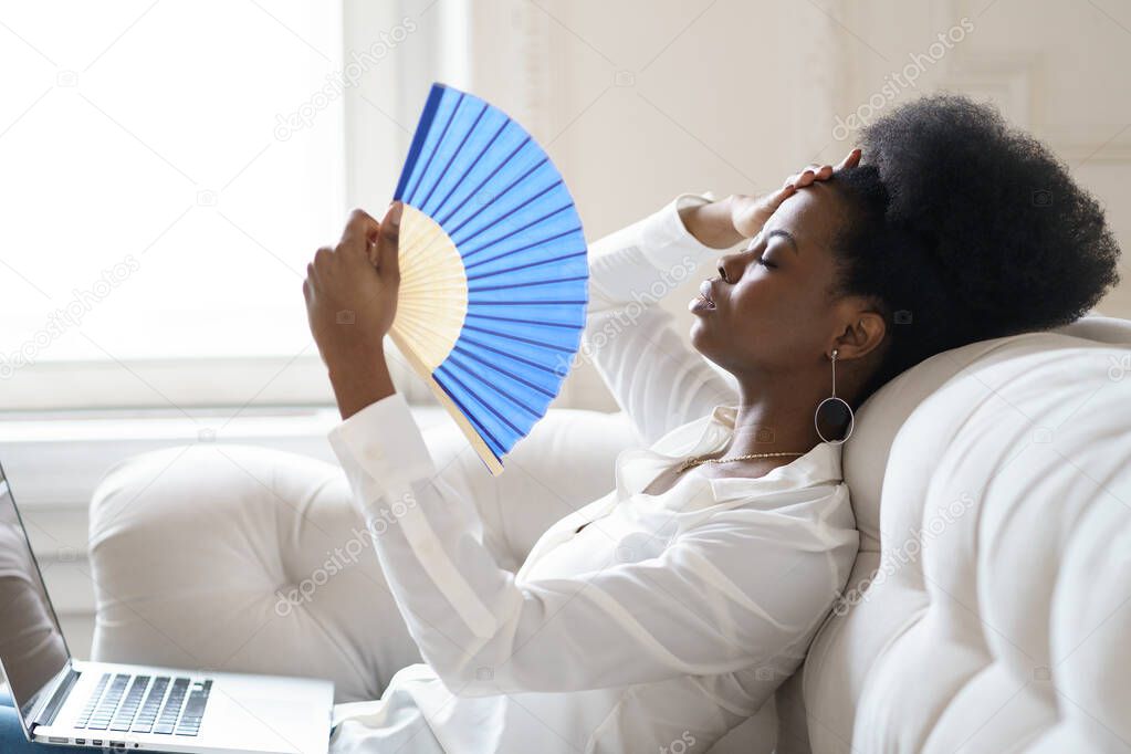 Afro business woman suffering from heat stroke sitting in living room at home using waving fan