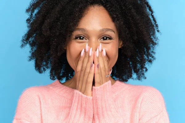 Szczęśliwa afrykańska kobieta z kręconymi włosami, uśmiechnięta, zakrywająca usta dłońmi, niebieskie tło. — Zdjęcie stockowe