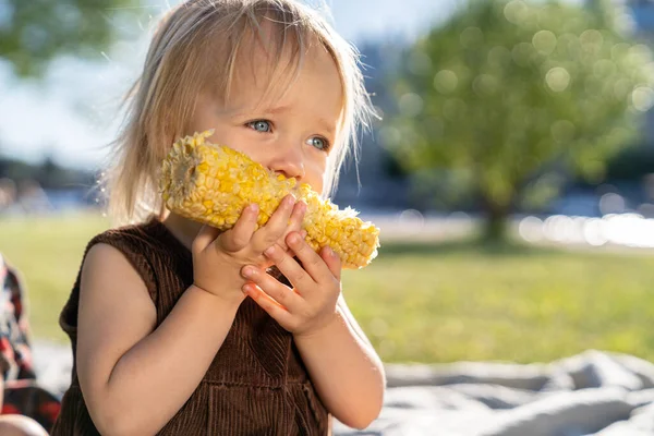 Küçük kız tatlı mısır koçanı yer, yaz günü çimenlerde ekoseye oturur. Sağlıklı beslenme. — Stok fotoğraf