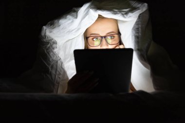 Woman in glasses lying in bed under the blanket using tablet late at night, can not sleep, addict news about Covid-19 or scrolling social networks. Insomnia, nomophobia, sleep disorder.   clipart
