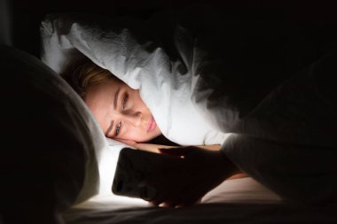 Woman lying in bed under the blanket using smartphone late at night, can not sleep, addict news about Covid-19 or scrolling social networks. Insomnia, nomophobia, sleep disorder.   clipart