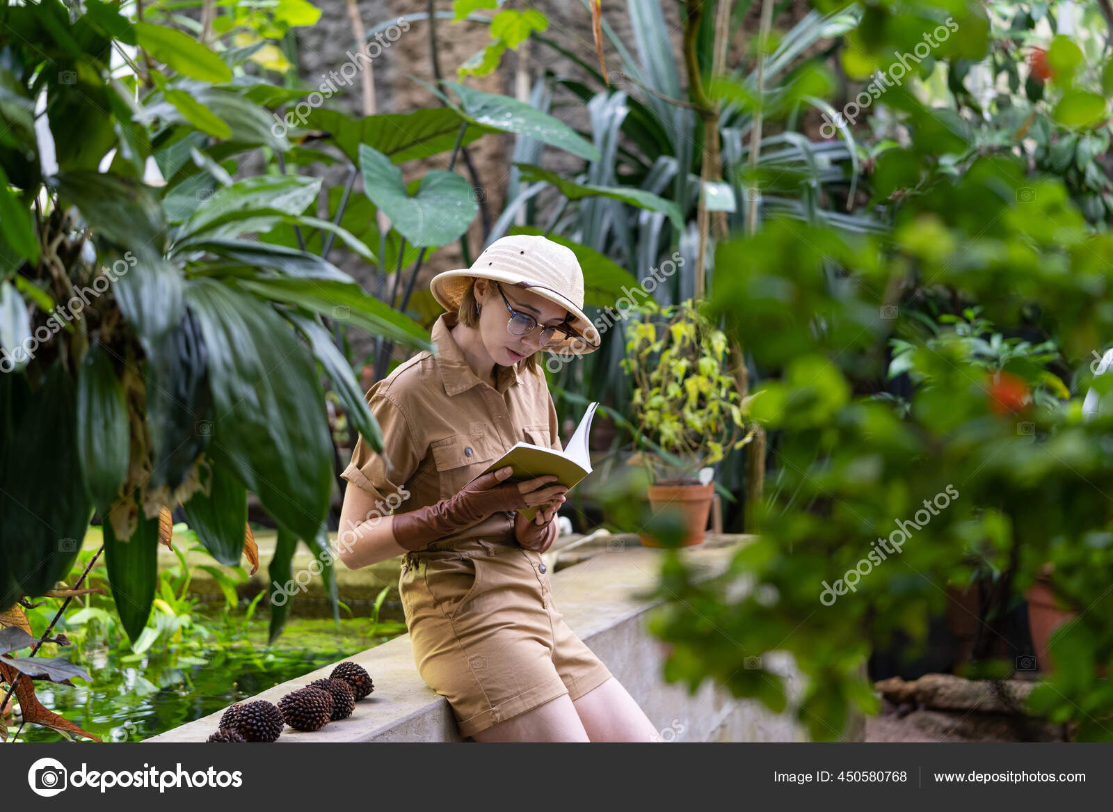 在温室里穿着狩猎风格衣服的女植物学家穿着帽子和卡其布衣服的自然主义者戴着树叶手套休息着阅读着四周环绕着植物的书籍
