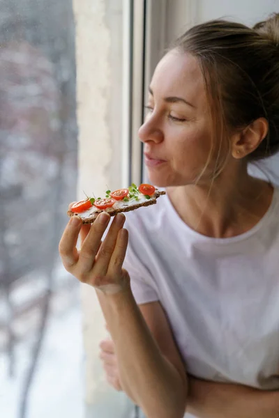 Egy nő rozsos, ropogós kenyeret eszik krémes vegetáriánus sajtos tofuval, paradicsommal, mikrozöldséggel. Egészséges élelmiszer — Stock Fotó