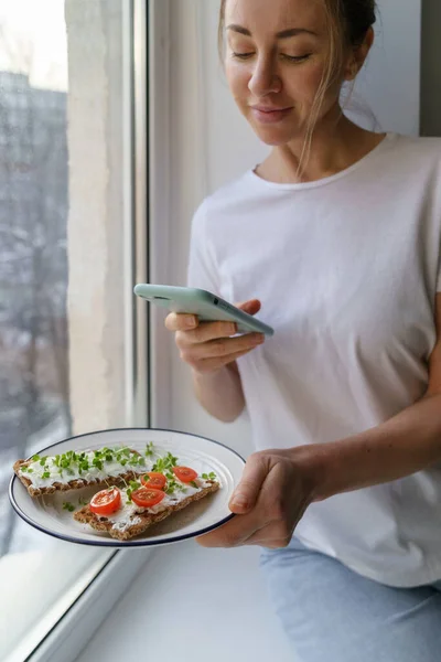 Femme prend une photo sur un téléphone intelligent, tenant la plaque avec du pain croustillant au seigle avec du fromage et des micro-verts — Photo