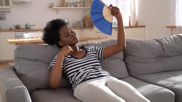 Μαύρη γυναίκα που υποφέρει από θερμοπληξία επίπεδη χωρίς air-condition κυματίζει ανεμιστήρα που βρίσκεται στον καναπέ στο σπίτι — Αρχείο Βίντεο