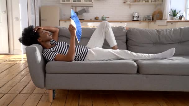 Zwarte vrouw die lijdt aan een hitteberoerte plat zonder airconditioner zwaaien ventilator liggend op de bank thuis — Stockvideo