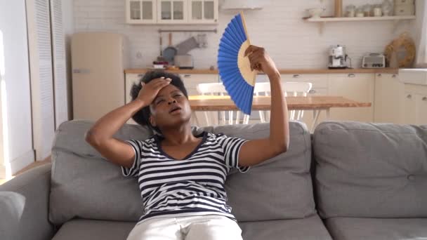Черная женщина страдает от теплового удара плоской без кондиционера размахивая вентилятором лежа на диване дома — стоковое видео