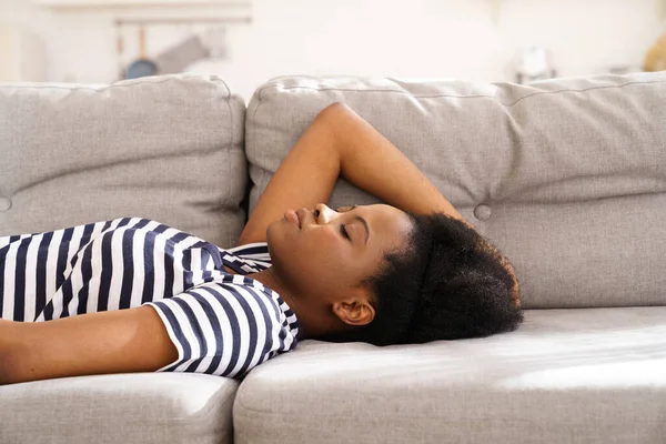 Negro joven mujer usar despojado camiseta descansando durmiendo en sofá en casa cerrado los ojos tomando un descanso — Foto de Stock