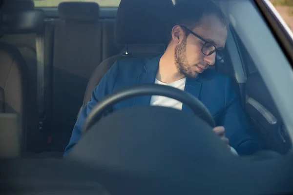 Tired business man falling asleep sitting inside her car seen through windshield, stopped to rest after driving. Exhausted, sleepiness, overworked driver concept.