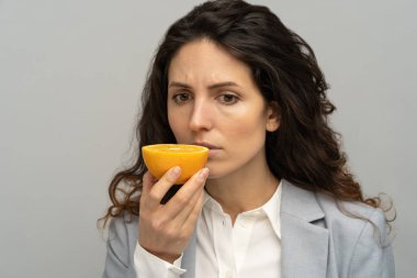 Sick business woman trying to sense smell of half fresh orange, has symptoms of Covid-19, corona virus infection - loss of smell and taste. One of the main signs of the disease. Studio grey background clipart
