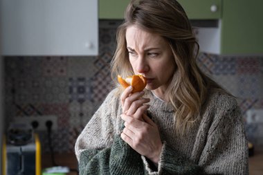 Sick woman trying to sense smell of fresh tangerine orange, has symptoms of Covid-19, corona virus infection - loss of smell and taste, standing at home. One of the main signs of the disease. clipart