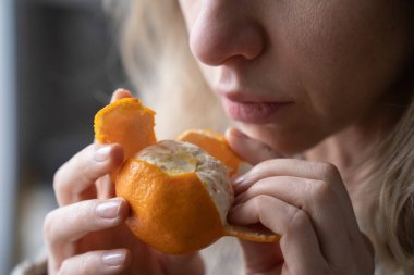 Sick woman trying to sense smell of fresh tangerine orange, has symptoms of Covid-19, corona virus infection - loss of smell and taste, standing at home. One of the main signs of the disease. clipart