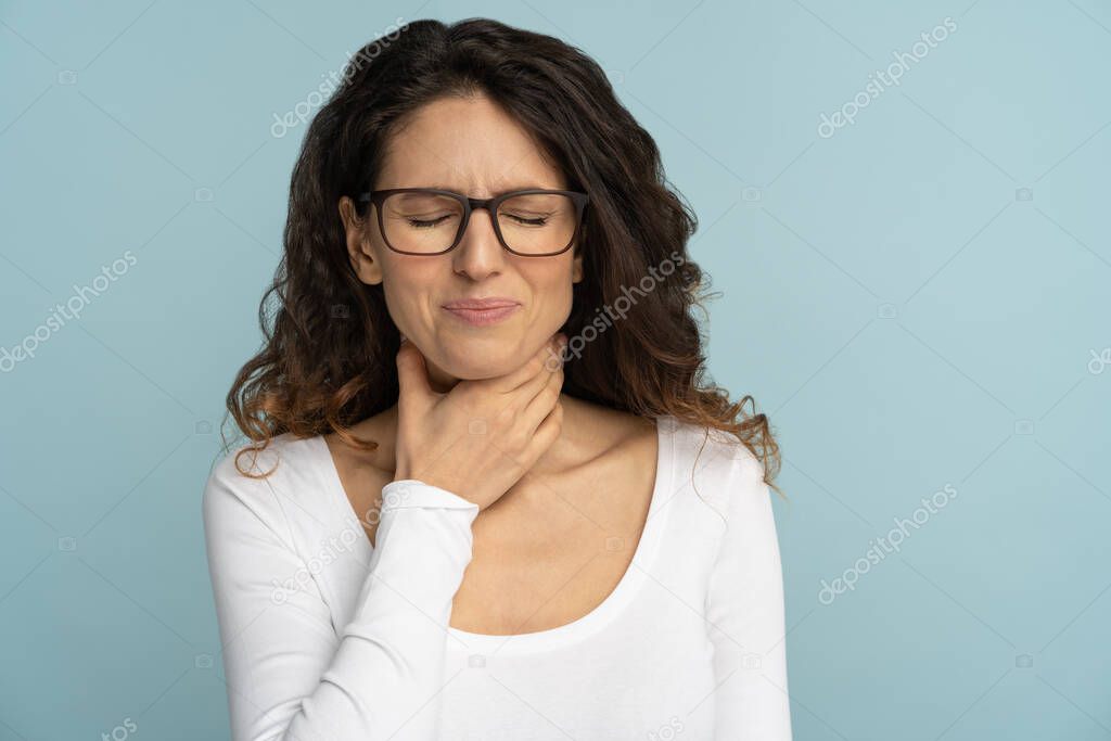 Sick woman having sore throat, tonsillitis, feeling sick, suffering from painful swallowing, angina, strong pain in throat, loss of voice, holding hand on her neck, isolated on studio blue background.