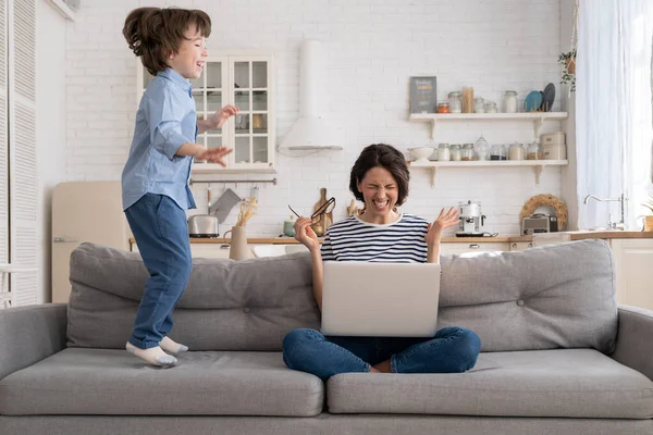 La mère fatiguée s'assoit sur le canapé, travaille sur un ordinateur portable à la maison, le petit enfant hyperactif saute en attirant l'attention. — Photo