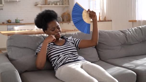 Mujer negra que sufre de golpe de calor plana sin aire acondicionado agitando ventilador acostado en el sofá en casa — Vídeos de Stock