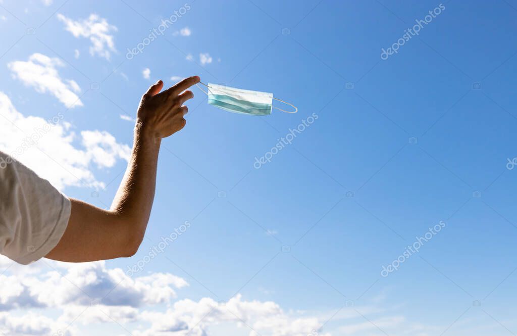 End of quarantine, covid-19. Man taking off and throw away used medical protective mask, holds it on her finger on blue sky background, enjoys life, breathes fresh air after coronavirus pandemic.