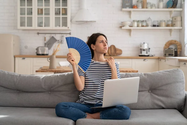 Mujer joven sufre de golpe de calor plana sin aire acondicionado agitando ventilador sentado en el sofá en casa —  Fotos de Stock